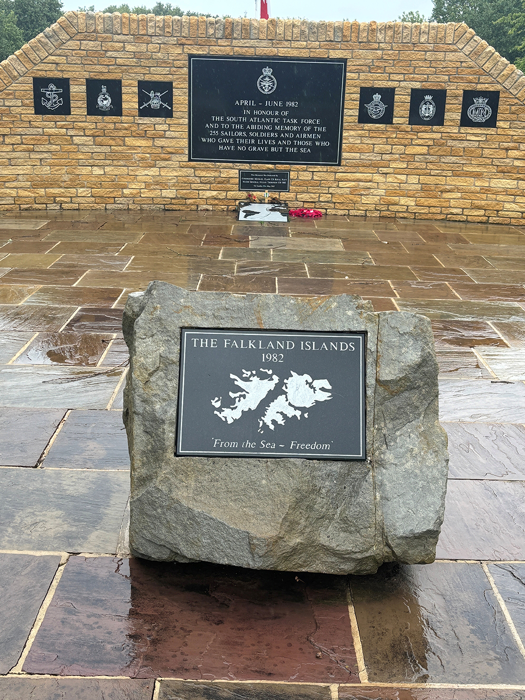 The Falkland Islands memorial stone, with inset plaque engraved with an image of the islands, with above, the words, in capital letters, 'The Falkland Islands 1982', and below, the words in title case, 'From the Sea ~ Freedom'. The stone is placed in front of a shaped brick wall, upon which, are placed further plaques, emblems and further dedications.